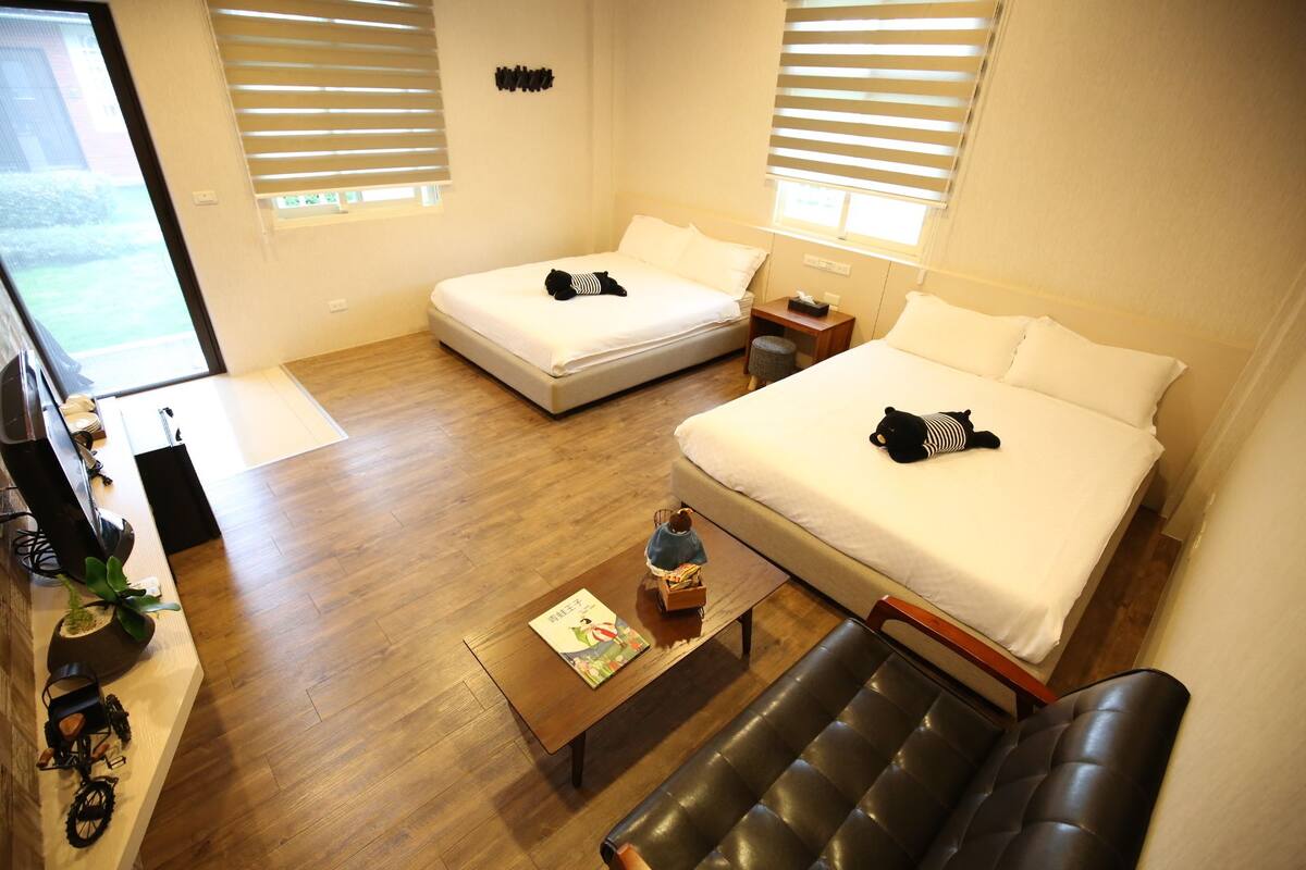 Quarto de hotel com duas camas de solteiro, uma cama de casal, uma mesa de centro de madeira com um livro, um banco e uma pequena mesa com um abajur.
