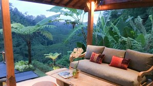 Un sofá de madera con cojines, una mesa de madera y dos otomanas en una habitación con vistas a exuberante vegetación.