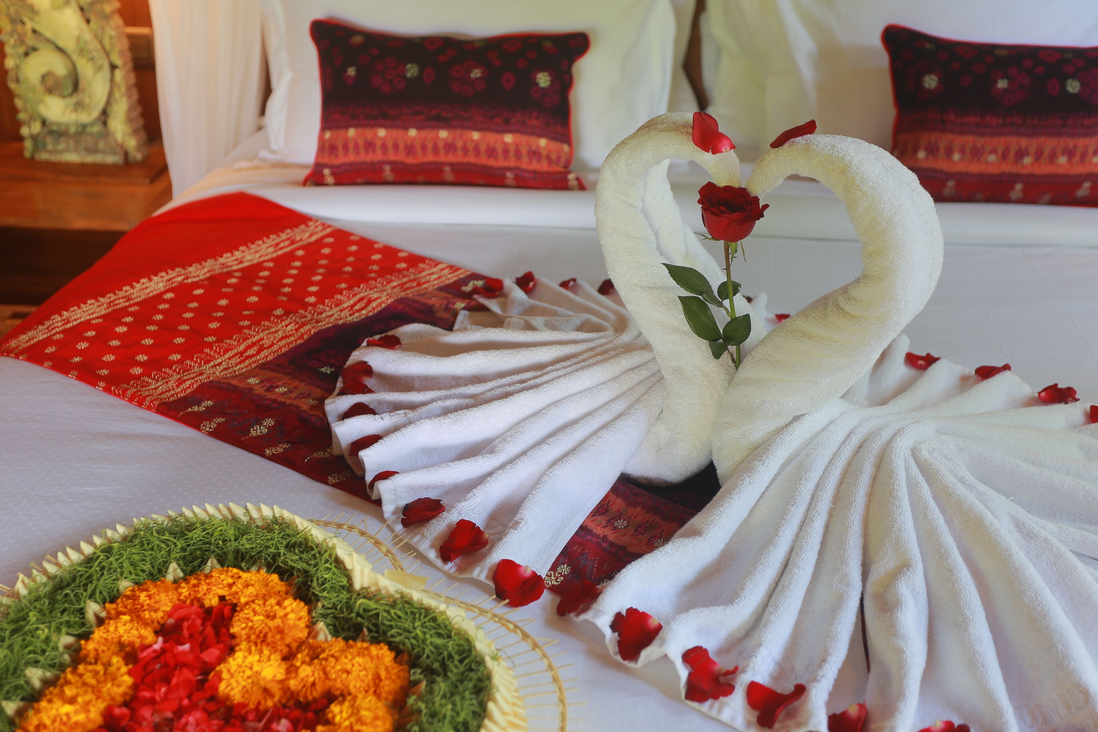 A bed with a white and red blanket, a bouquet of flowers, and a swan-shaped towel with rose petals.