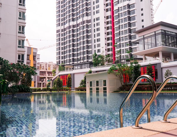 A modern pool area with a clear pool, surrounded by buildings, including a high-rise with balconies and a smaller structure with red accents.