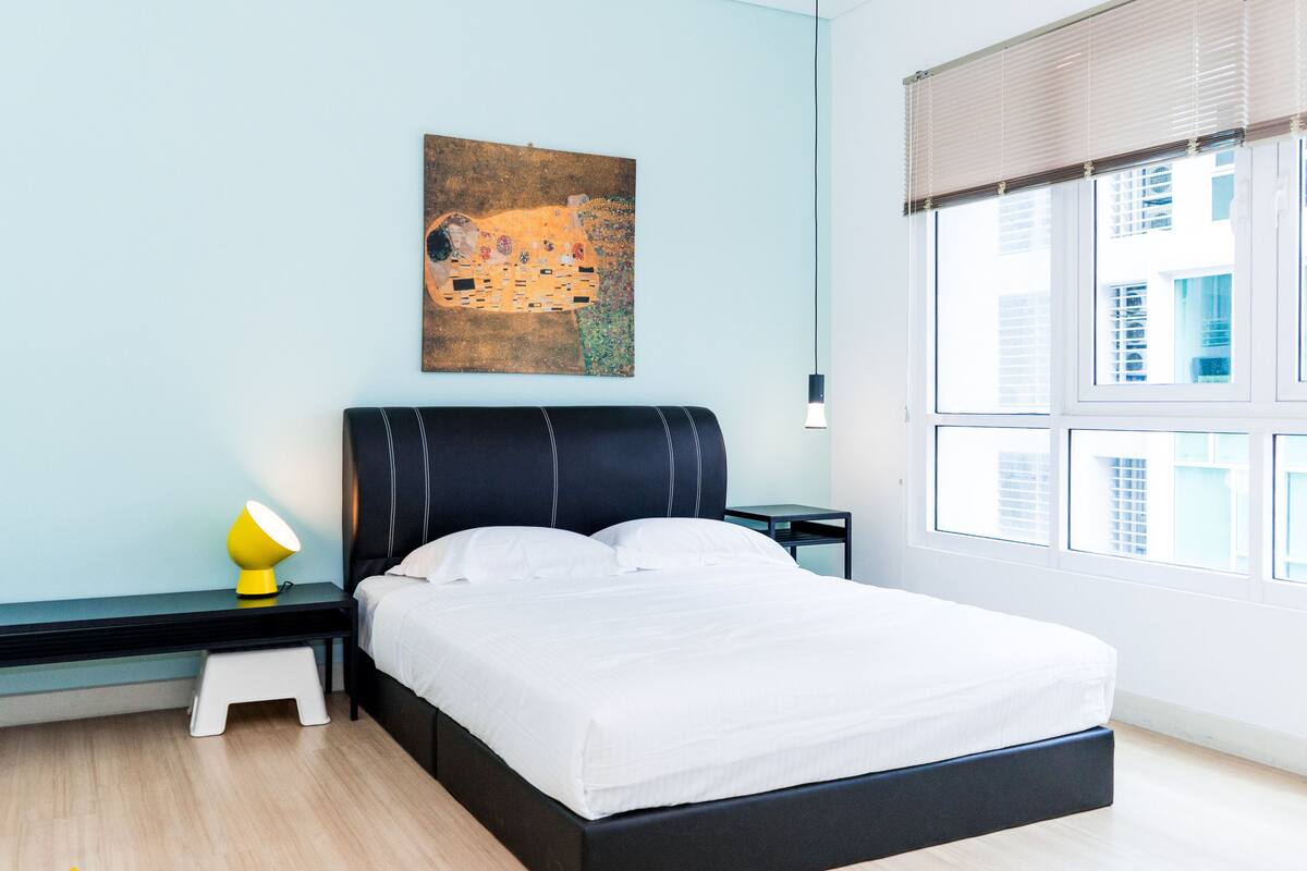 Une chambre moderne avec une tête de lit noire, du linge de lit blanc, une petite table de chevet avec une lampe et une fenêtre avec des stores.
