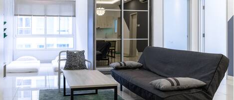 A modern living room with a grey sofa, a wooden coffee table, and a white armchair. There is a large window with blinds, a green rug, and a view of the interior space.
