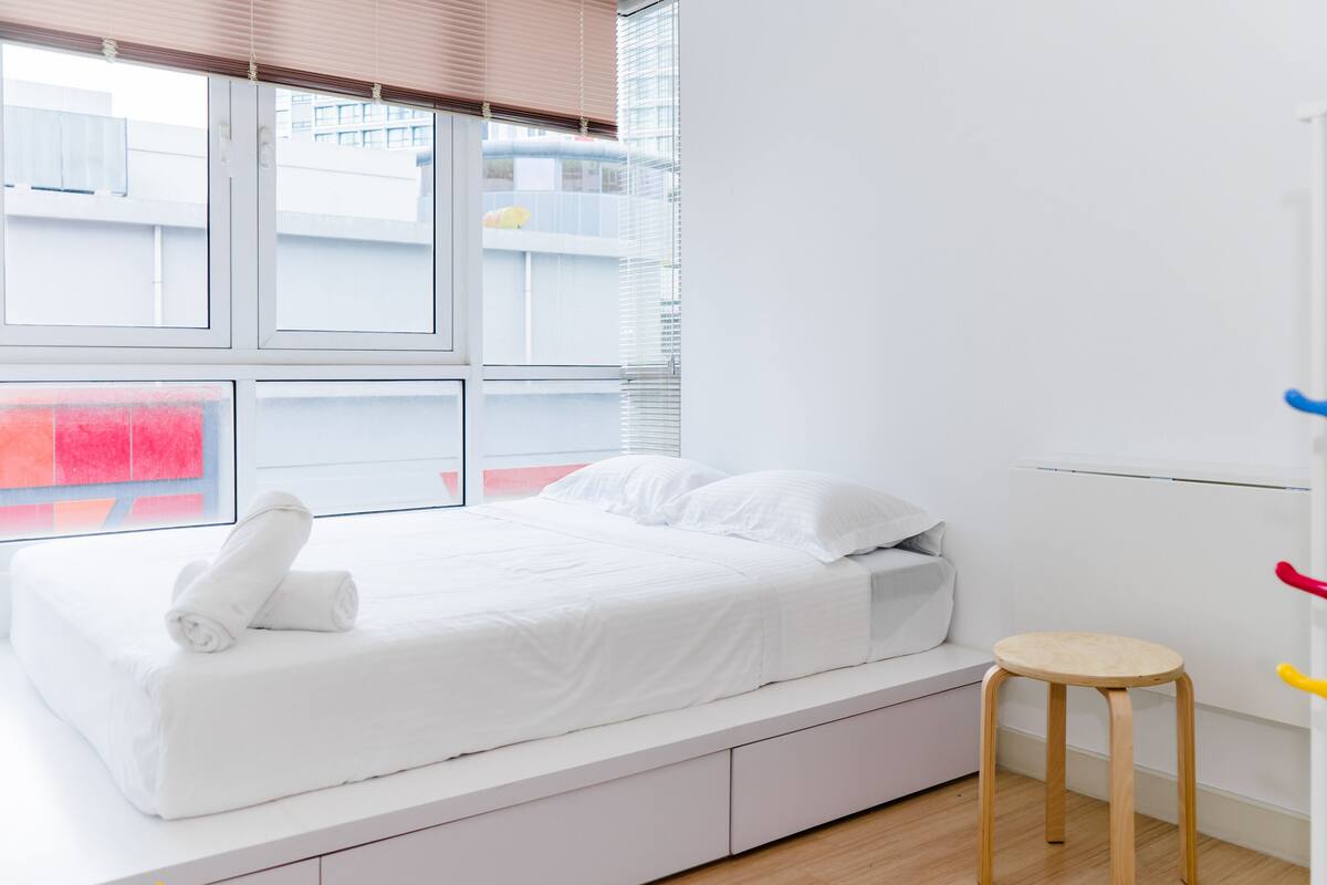 Une chambre à coucher au style minimaliste, avec un lit, un petit tabouret et une fenêtre équipée de stores.