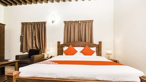 A neatly made bed with wooden headboard, two orange pillows, and a white bedspread. There is a nightstand with a lamp, a chair, and a wooden desk with a lamp.