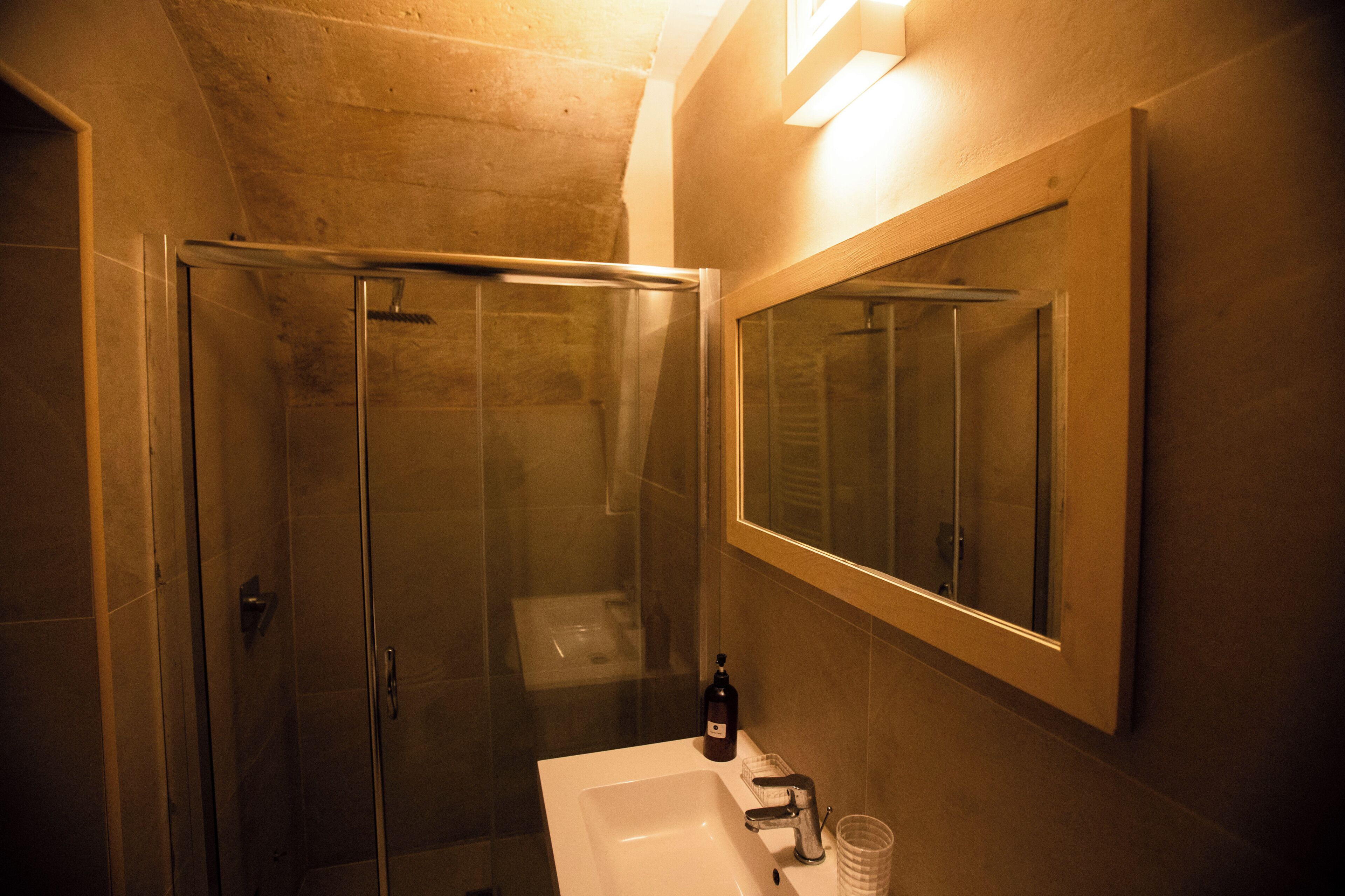 Bagno con cabina doccia in vetro, lavandino bianco, uno specchio e una bottiglia di sapone.