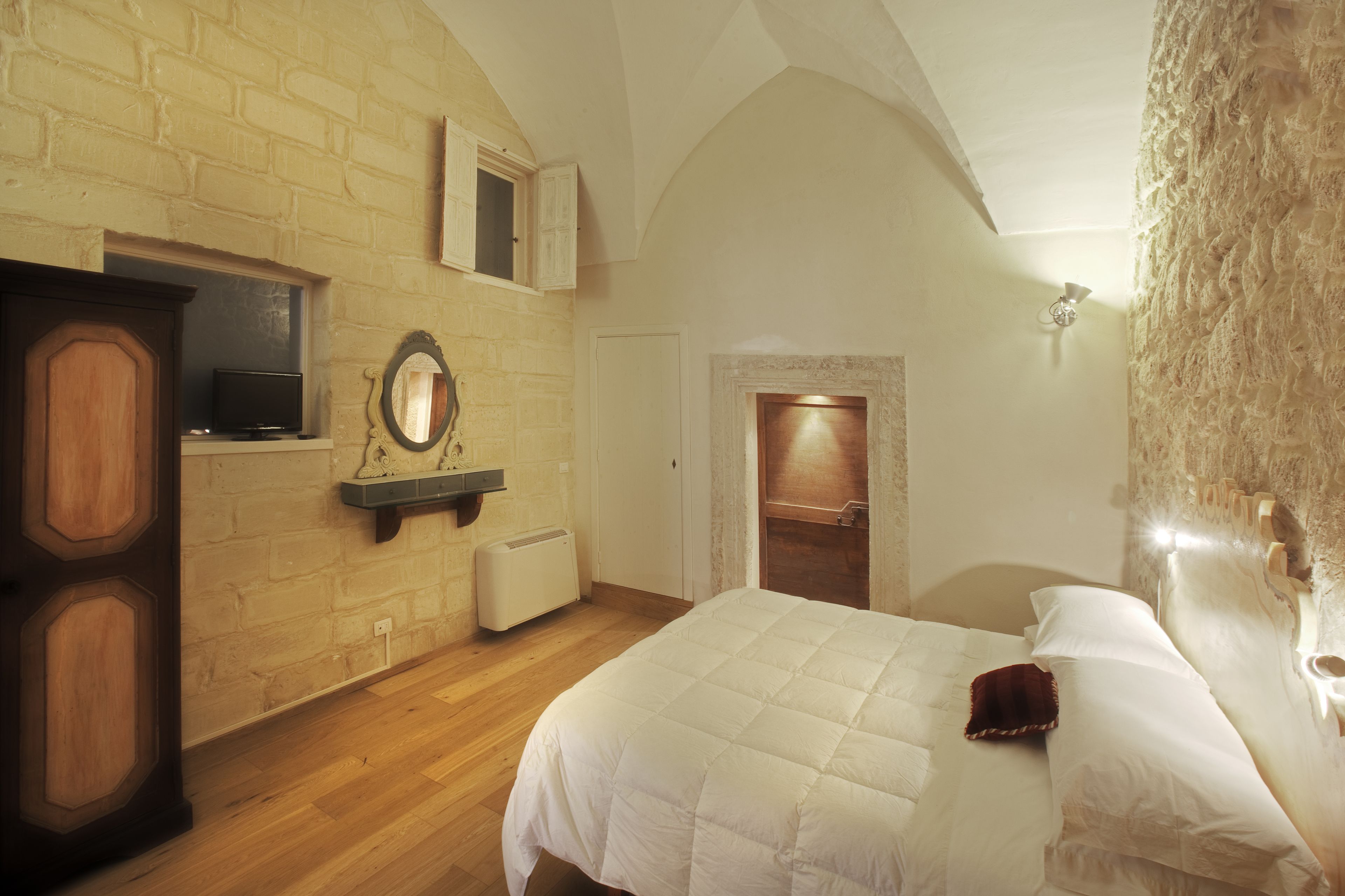 A bedroom with a wooden floor, a bed with white bedding, a wooden wardrobe, a mirror, and a small television on a shelf.