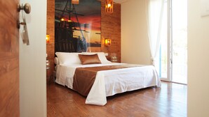 A bedroom with a wooden floor, a bed with white bedding and a brown throw, a large window with curtains, and a sunset mural on the wall.
