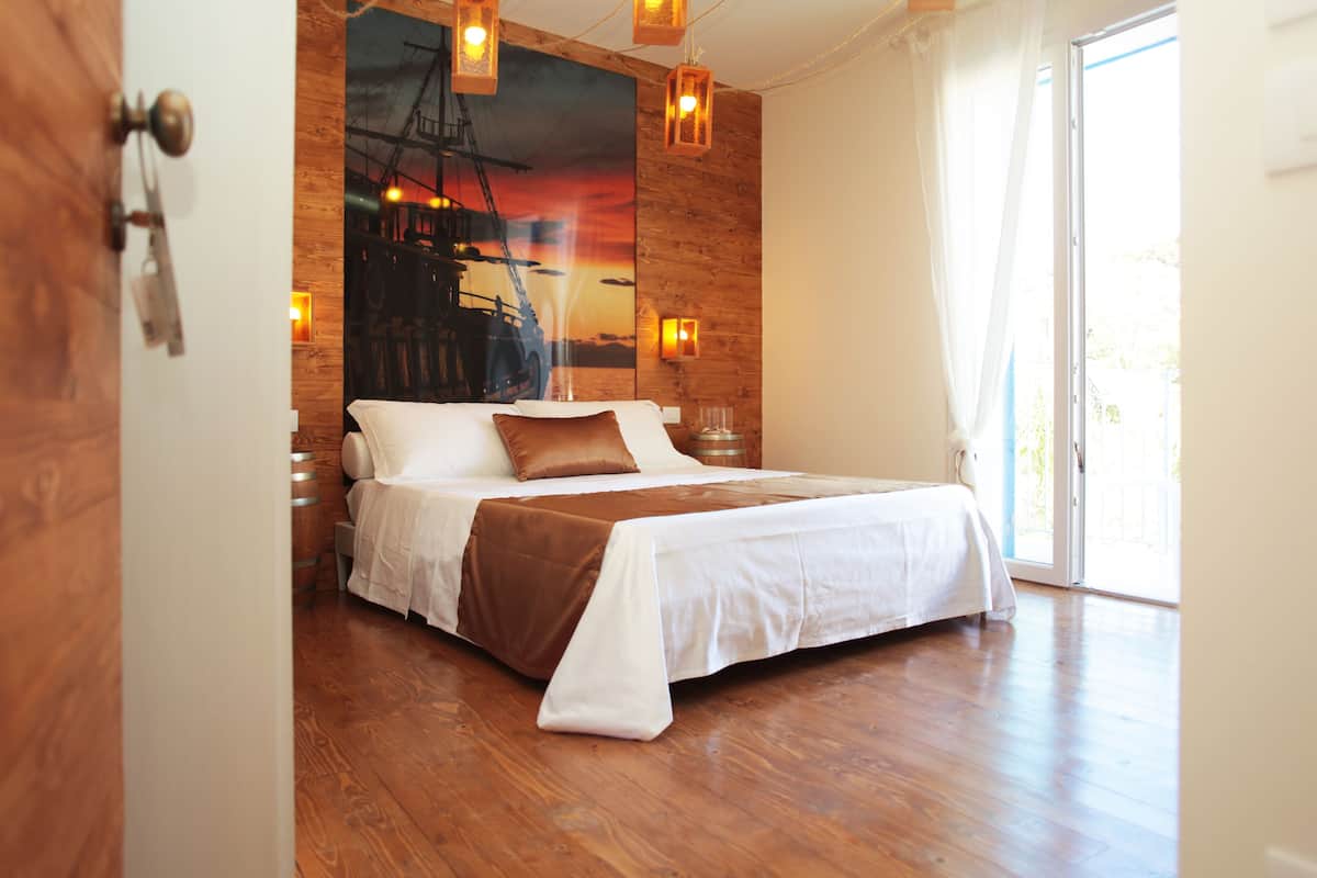 Une chambre avec un plancher en bois, un lit recouvert de draps blancs et d’un plaid marron, une grande fenêtre avec des rideaux et une fresque représentant un coucher de soleil sur le mur.