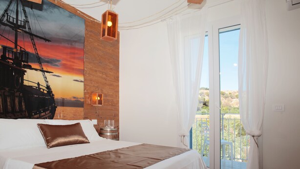 A bedroom with a large bed, a wooden accent wall, a painting of a ship, and a view through a glass door.