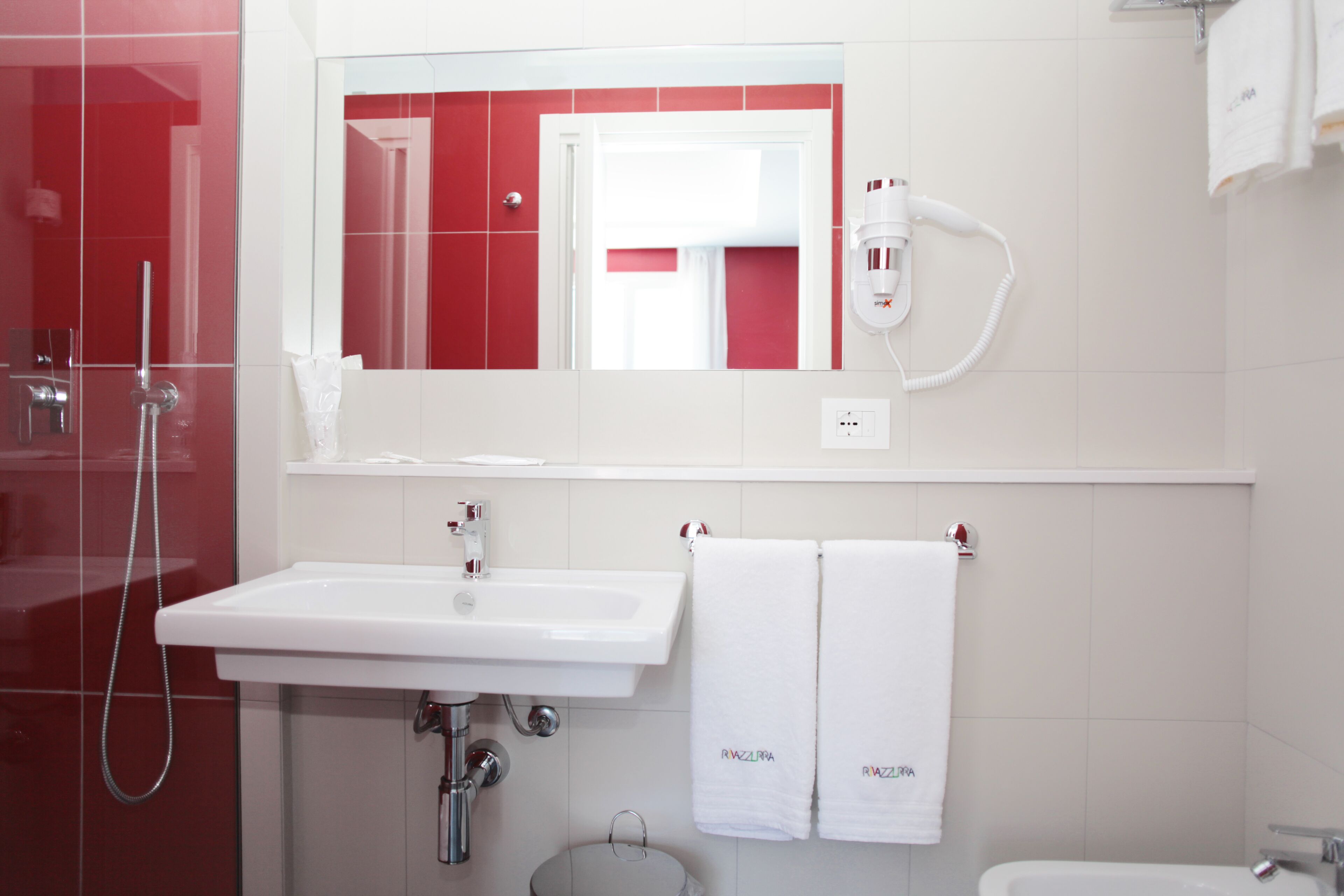 Ein Badezimmer mit einem weißen Waschbecken, einem Spiegel, einer Dusche und zwei weißen Handtüchern, die auf einem Ablagegestell hängen.
