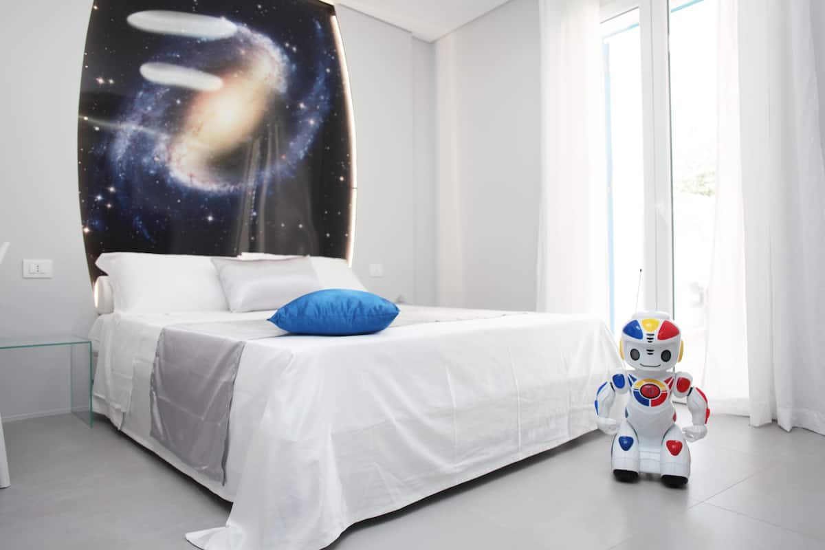 Une chambre à coucher avec un lit, un oreiller bleu, un jouet robot et un mur décoré sur le thème de la galaxie.