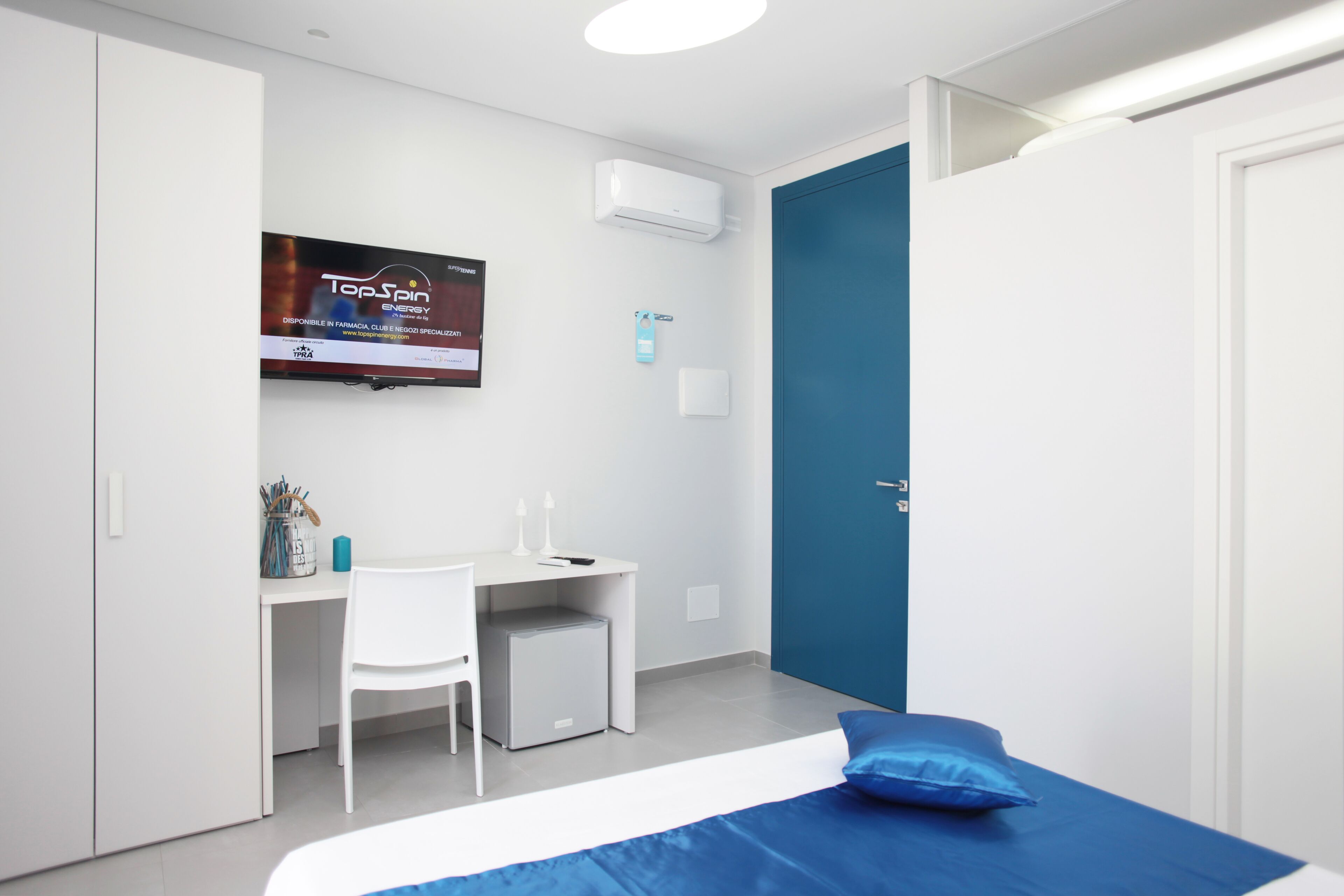 A modern hotel room with a bed, a desk, a chair, a TV mounted on the wall, and a door.