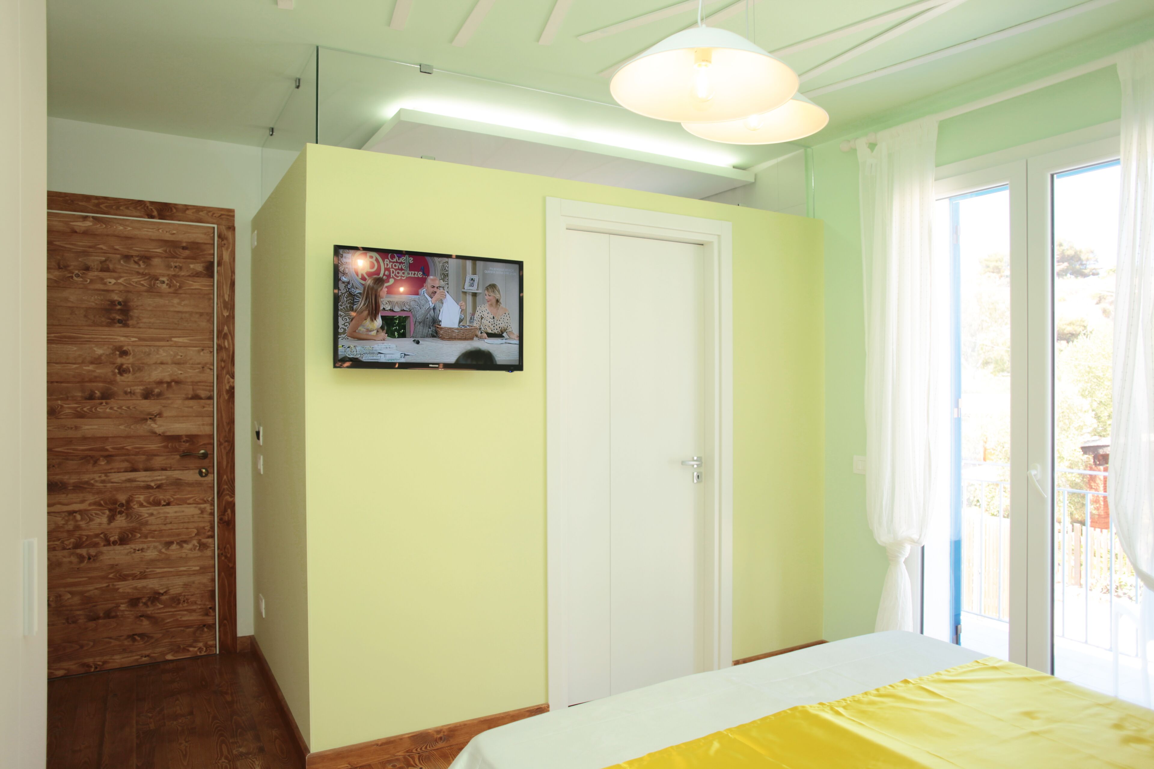 A bedroom with a bed, a wooden door, a white wall, and a television on the wall.