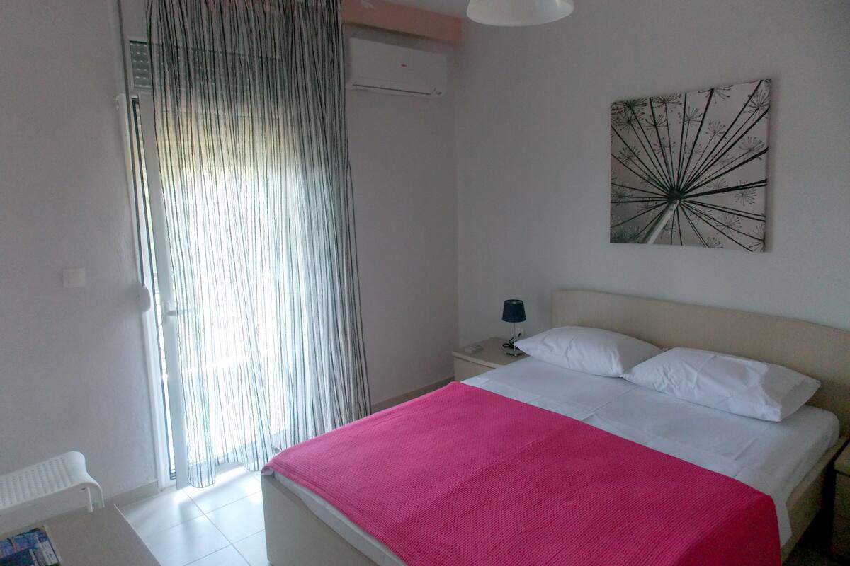 A bedroom with a bed, a nightstand, a lamp, a window with curtains, and a wall-mounted artwork.