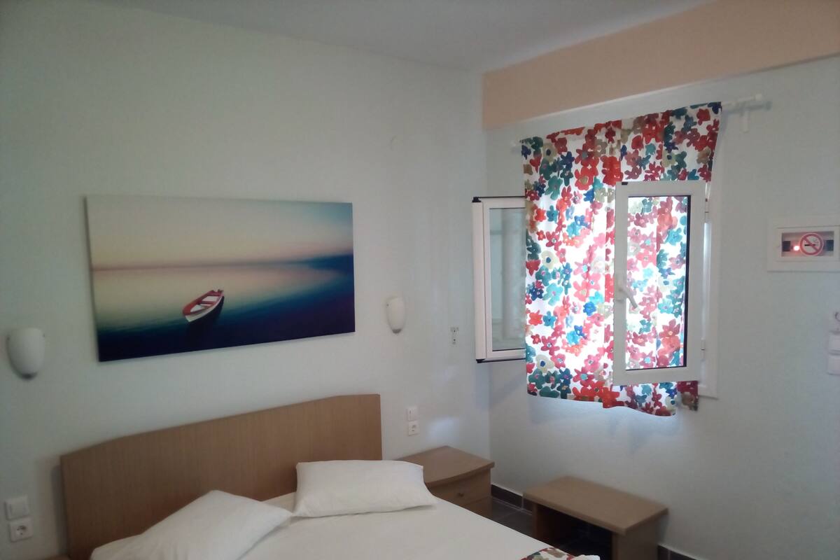 A hotel room with a bed, a nightstand, a small table, a window with floral curtains, and a painting on the wall.