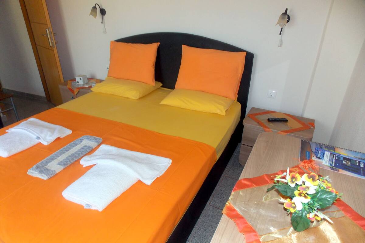 A hotel room with a bed, pillows, towels, a nightstand, a remote control, and a decorative flower arrangement.