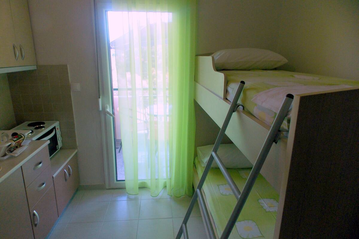 A small, compact room with a bunk bed, a kitchenette with a microwave, and a sliding glass door with sheer curtains.
