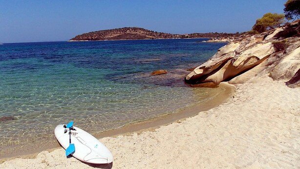 A clear beach with a surfboard and paddleboard on the sand, a rocky shoreline, and a distant island. - Nikos Apartments (Sithonia)