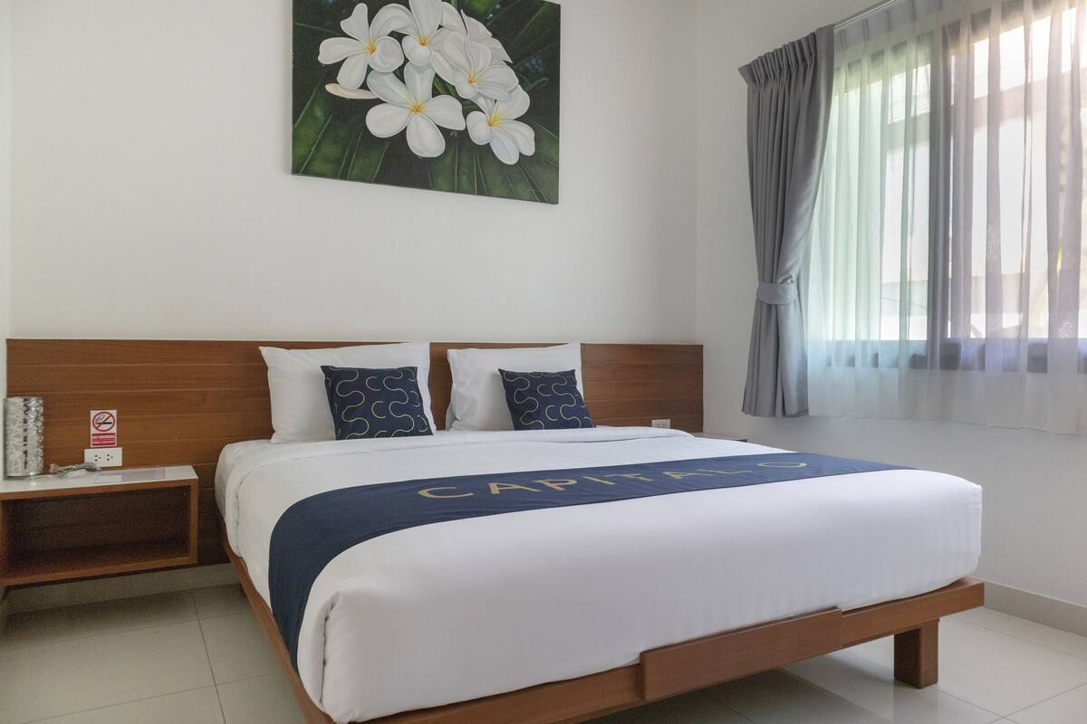 Een netjes opgemaakt bed met een wit-blauwe spreideken, twee kussens, een nachtkastje met een boek en een telefoon, en een schilderij van witte bloemen aan de muur.