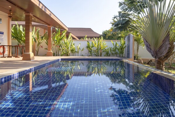 A swimming pool with blue tiles, surrounded by plants and a building with a tiled roof.