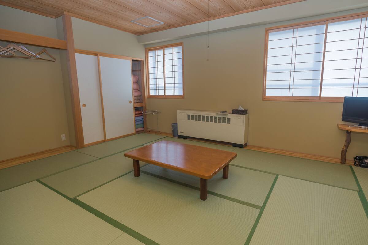Une pièce de style japonais traditionnel, avec un sol recouvert de tatamis, une table basse, des portes coulissantes, un radiateur et un téléviseur posé sur un meuble en bois.