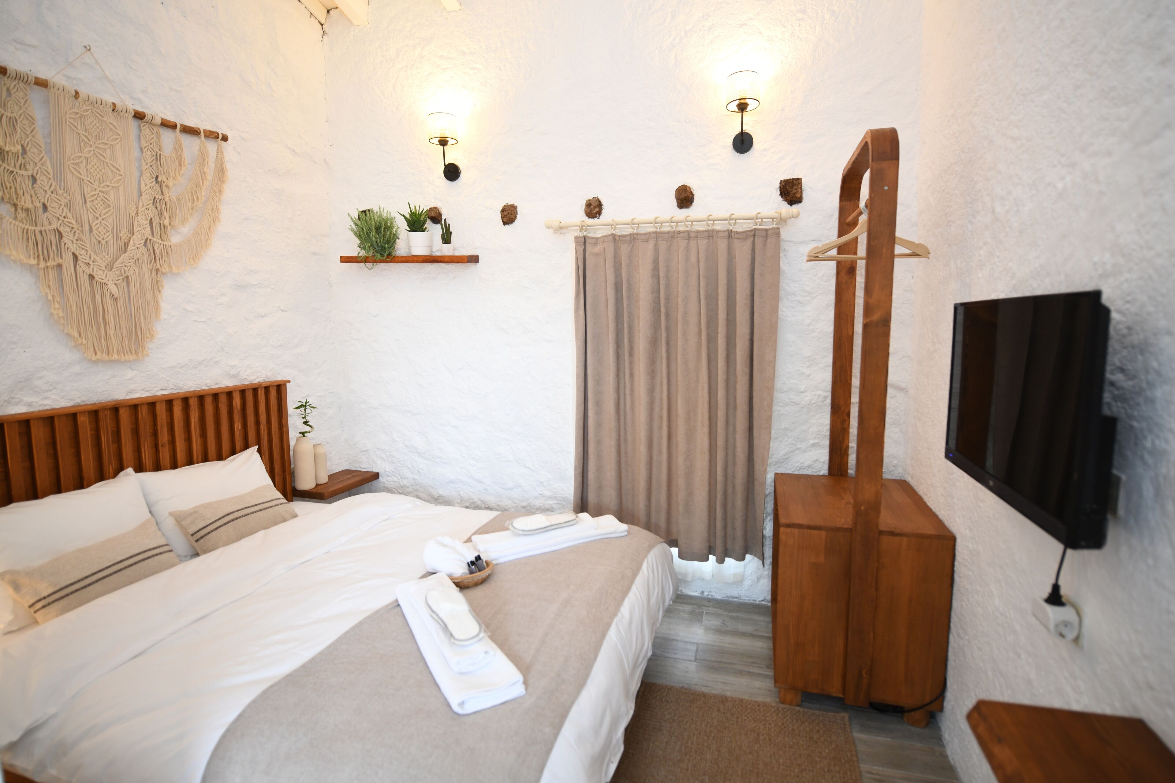 Une chambre à coucher bien aménagée, avec un lit en bois, une table de chevet agrémentée d’une plante, une décoration murale en macramé suspendue, une chaise en bois et un téléviseur fixé au mur.