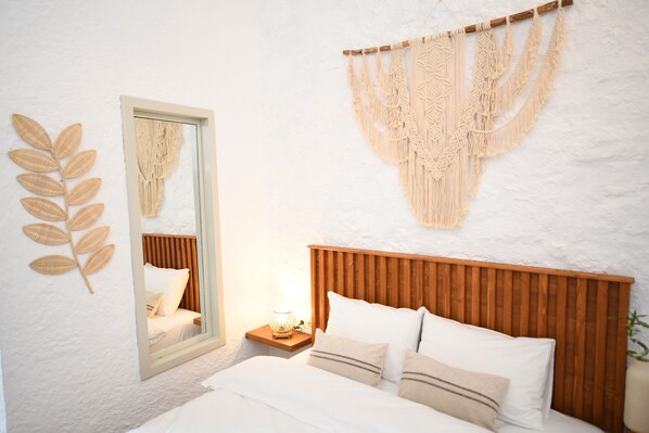 A bedroom with a wooden headboard, a bed with white bedding, a nightstand with a lamp, a mirror, and macramé wall art.