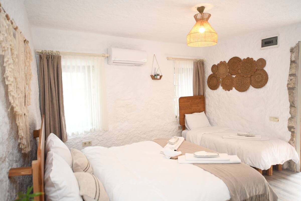 A room with two beds, a hanging light fixture, a wall-mounted air conditioner, and a window with curtains.