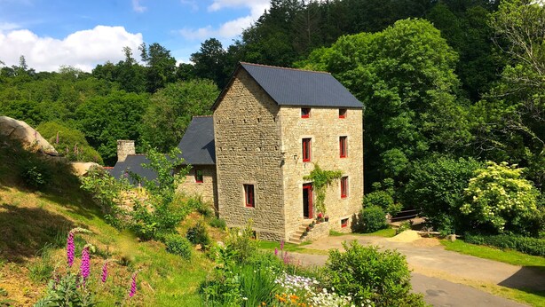 A stone building with a sloped roof, surrounded by lush greenery and a variety of colorful flowers. - Authentic mill with feet in water (near the sea) (Le Vieux-Marché)