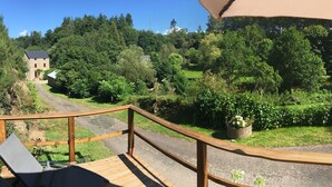 A wooden deck with a view of a stone building surrounded by lush greenery and a clear sky. - Authentic mill with feet in water (near the sea) (Le Vieux-Marché)