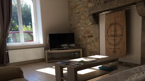 A living room with a stone wall, a television on a wooden stand, a coffee table with books, and a window with curtains. - Authentic mill with feet in water (near the sea) (Le Vieux-Marché)