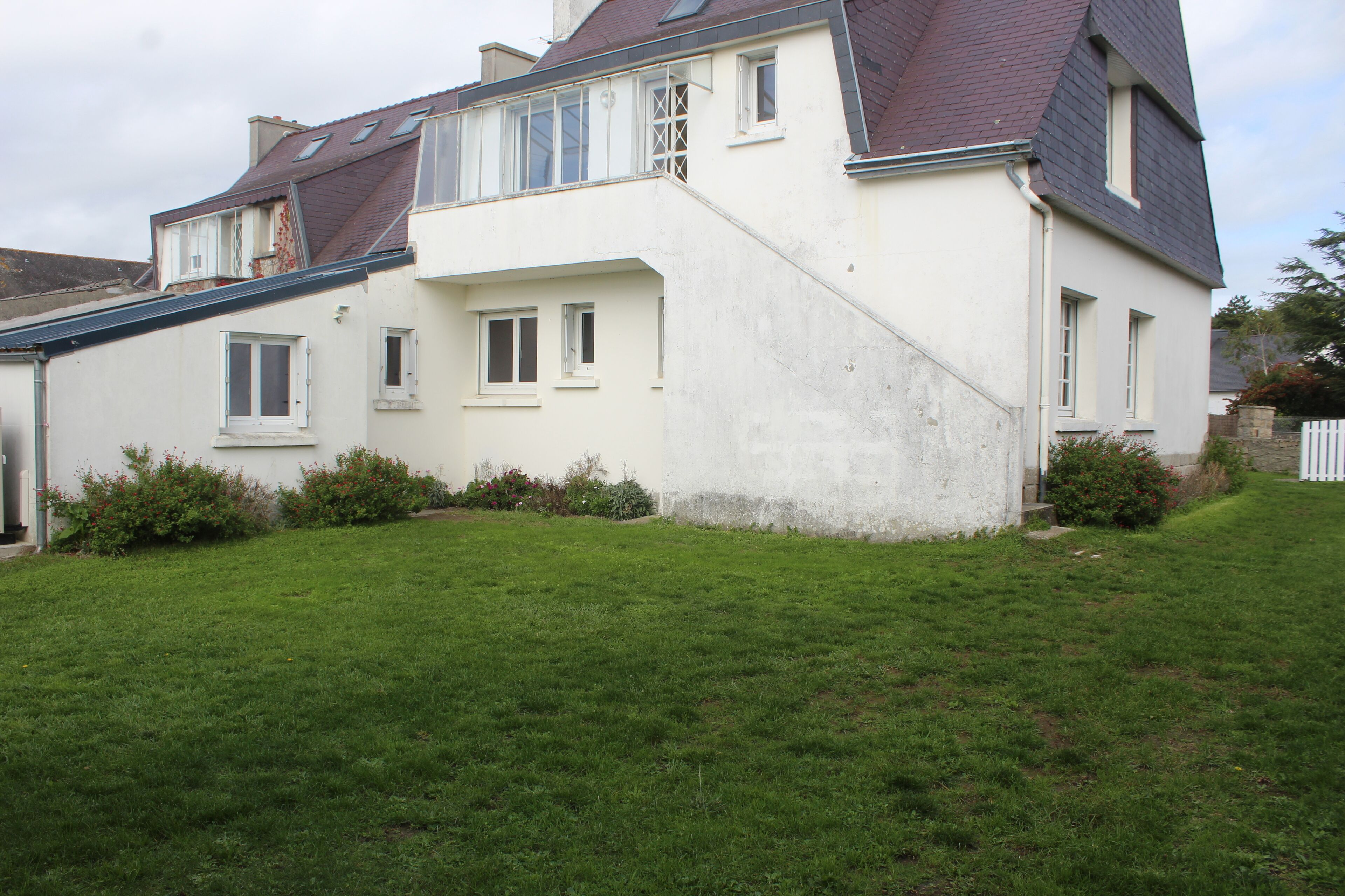 Une maison à deux étages, avec un toit en pente, des murs extérieurs blancs et un petit jardin devant, avec de la pelouse et des arbustes.