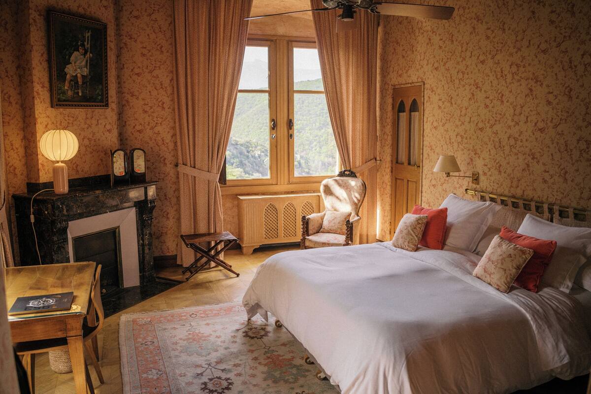 Un dormitorio con cama, una silla, una chimenea y una ventana con cortinas.