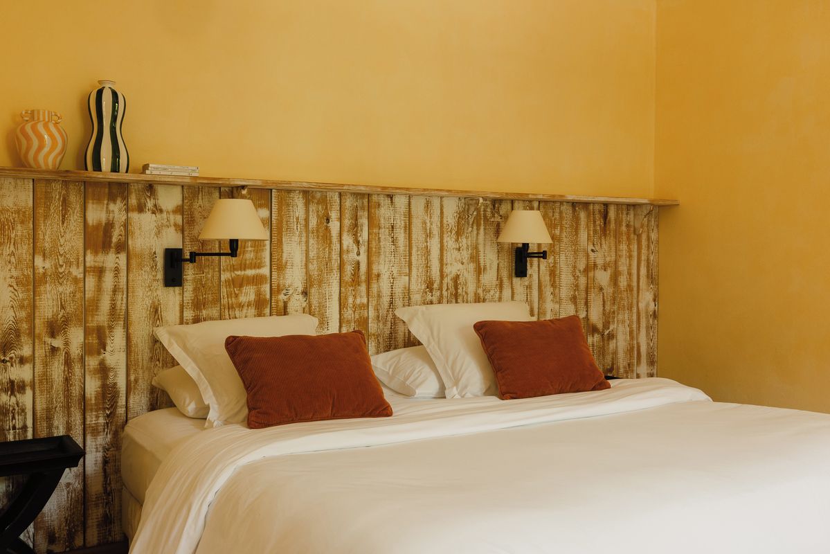 Quarto de hotel com cabeceira de madeira, duas luminárias de apoio, cama com roupa de cama branca e almofadas castanhas, e mesa de cabeceira com abajur.