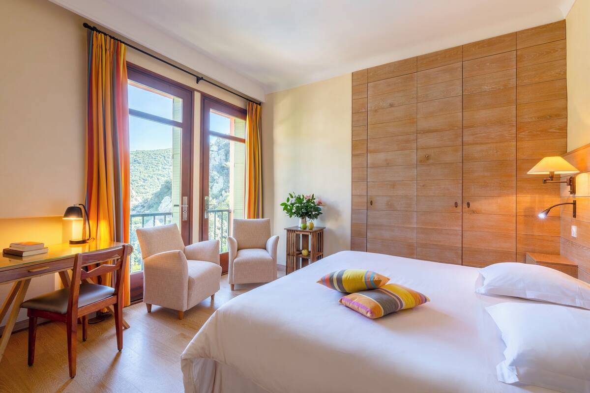 Habitación de hotel con una cama grande, dos sillones, un escritorio con lámpara, una mesita con una planta y un armario de madera.