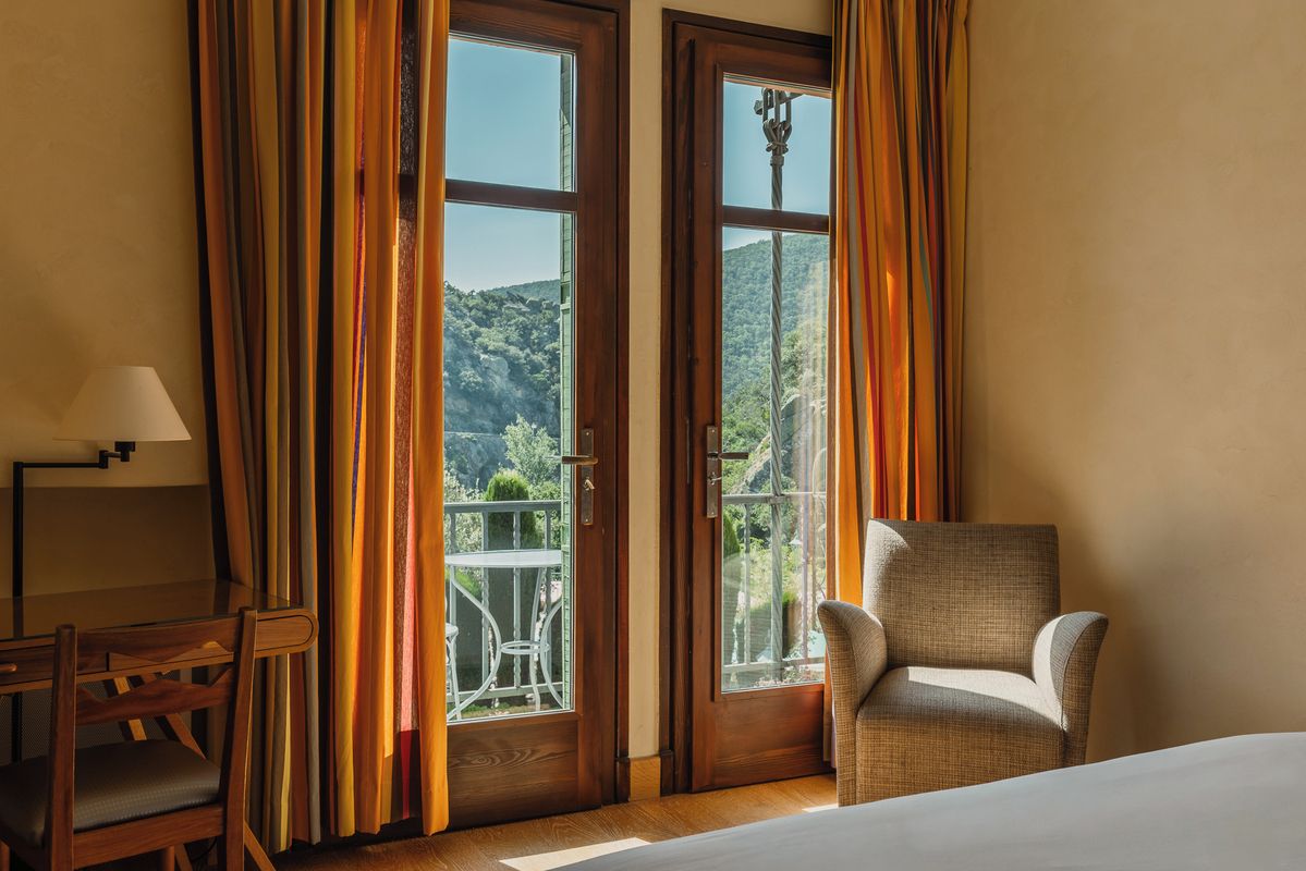 A room with a bed, a chair, and two doors leading to an outdoor area with a view of a hill.