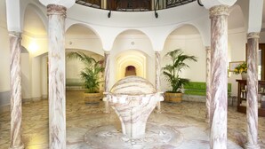 Un vaste hall d’entrée orné de colonnes en marbre, une fontaine centrale et un escalier menant à un balcon.