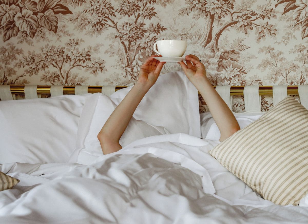 A person holding a white cup above a bed with white bedding and floral wallpaper.