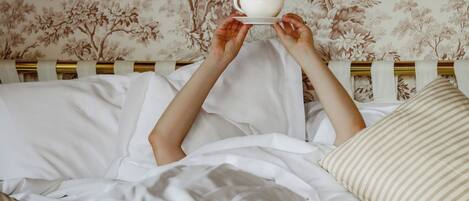 A person holding a white cup above a bed with white bedding and floral wallpaper.