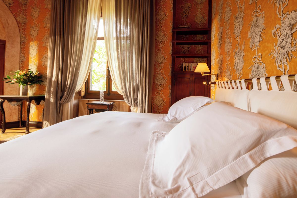 A hotel room with a large bed, a bedside table with a lamp, a window with curtains, and a bookshelf.