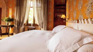 A hotel room with a large bed, a bedside table with a lamp, a window with curtains, and a bookshelf.