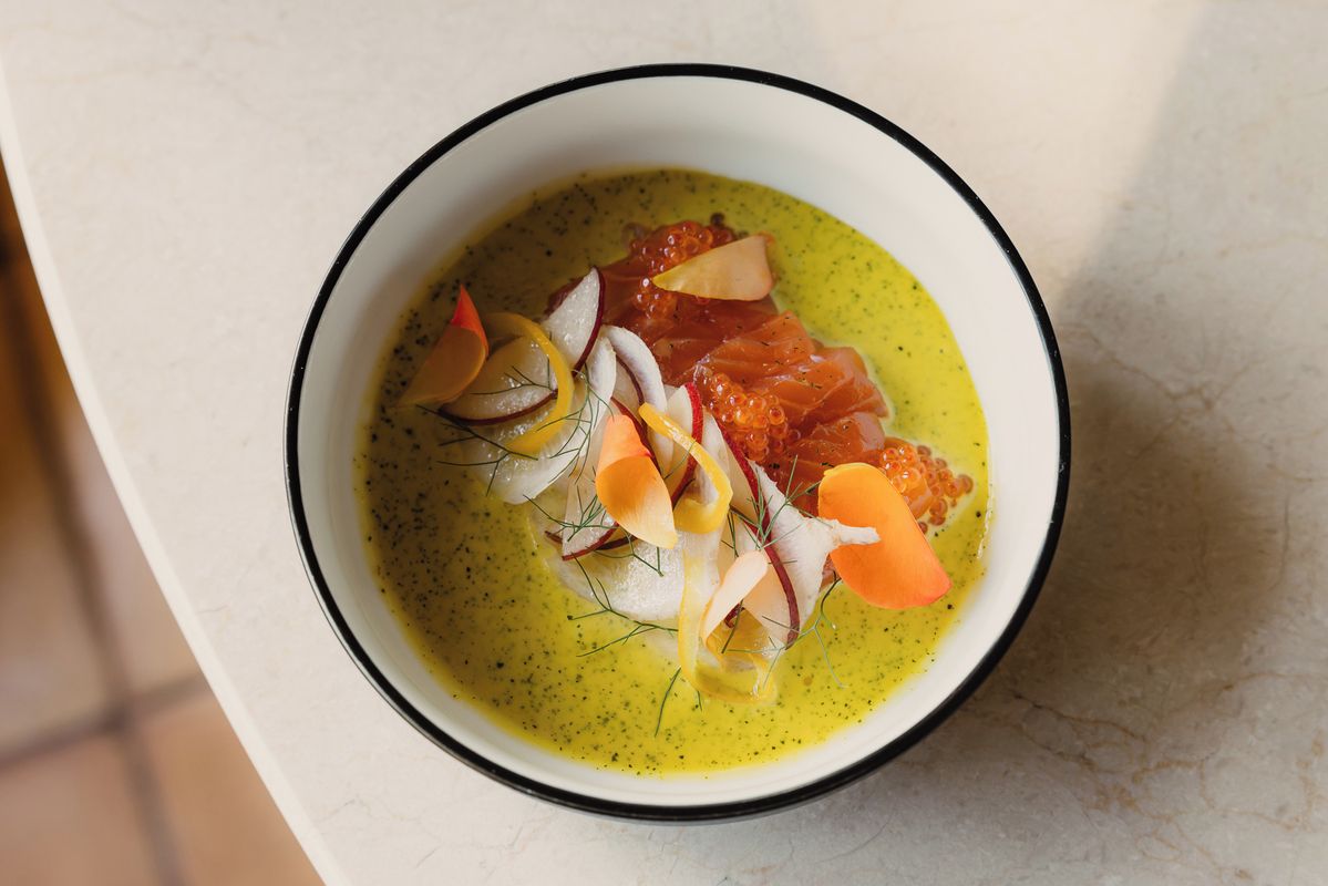 A bowl of food with a green sauce, fish, and garnishes on a white surface.