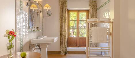 Un baño con lavamanos, espejo y toallero. Hay una ventana con cortinas, y una mesa con un florero y una manzana.