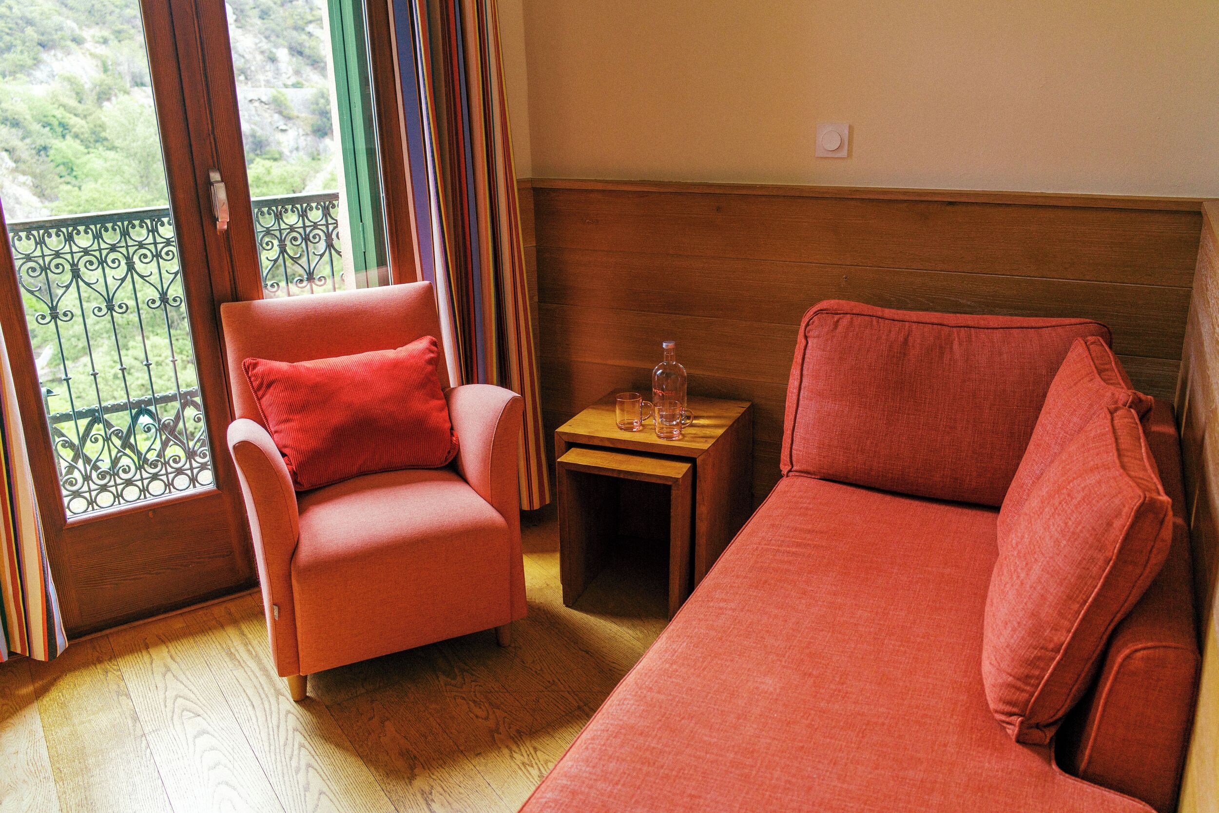 A room with a wooden floor, a red sofa, a red armchair, a small wooden table with a bottle and glasses, and a balcony with a wrought iron railing.