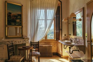 A bathroom with a marble sink, a wooden chair, a radiator, and a large window with curtains. - Le Grand Hôtel (Molitg-les-Bains)