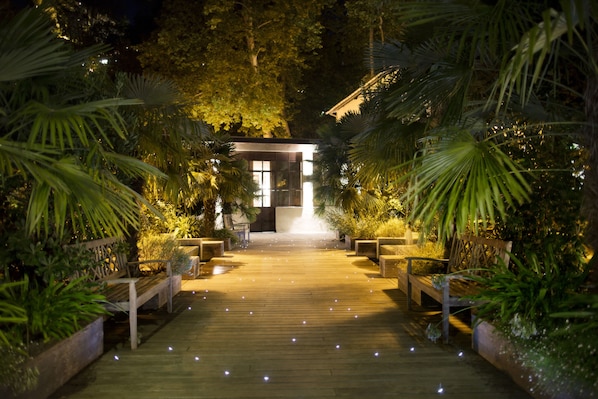 A wooden pathway leading to a building with a glass facade, surrounded by lush greenery and benches. - Le Grand Hôtel (Molitg-les-Bains)