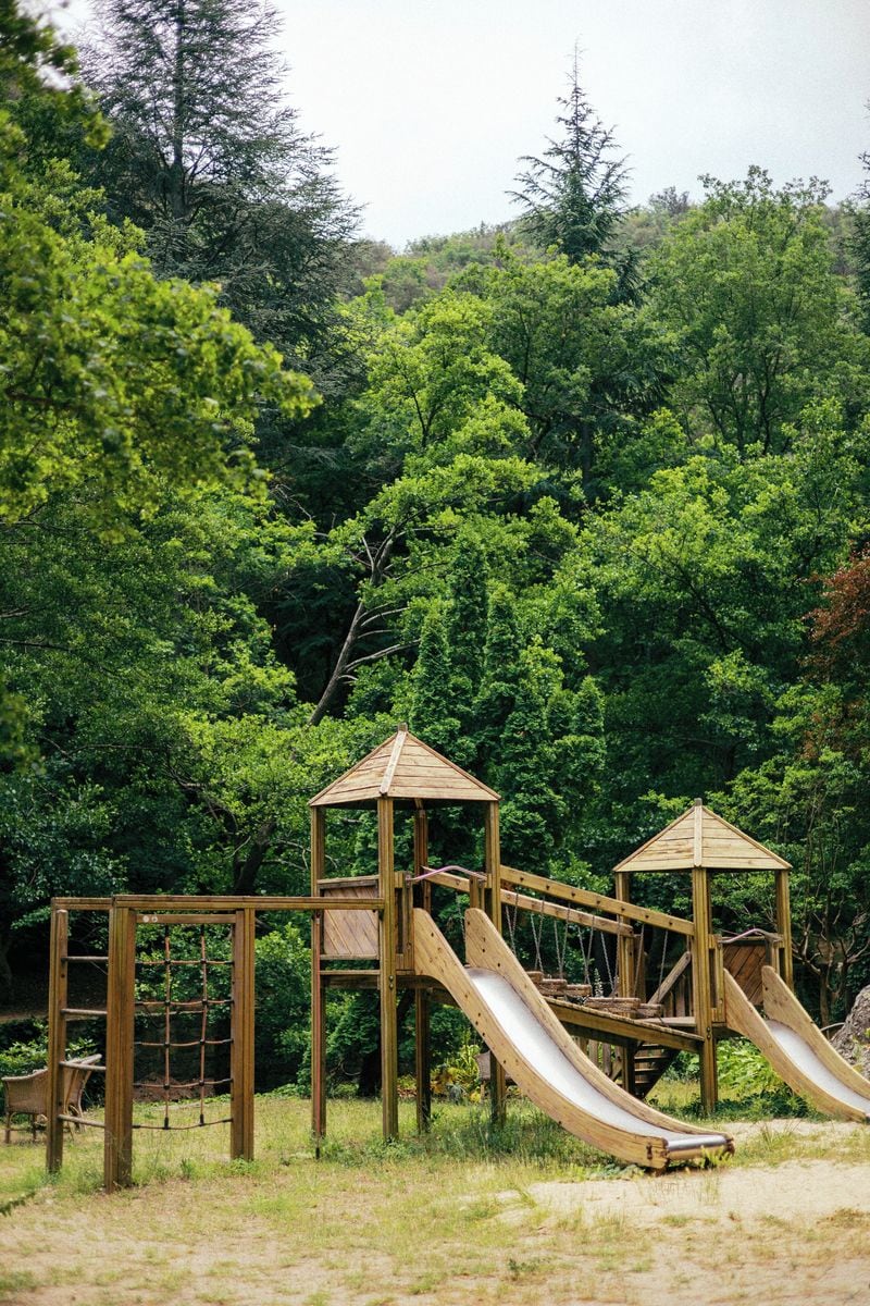 A playground with wooden slides and climbing structures, surrounded by dense greenery.
