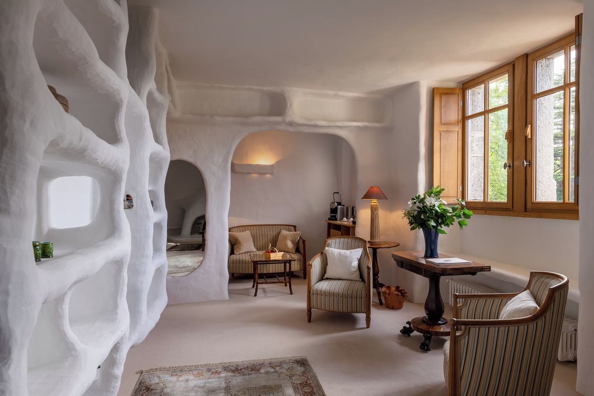 Una habitación con un diseño de pared blanco y único, similar a una cueva, un rincón de descanso acogedor con sofá y sillones, una mesita con una lámpara, y una ventana con vistas a zonas verdes.