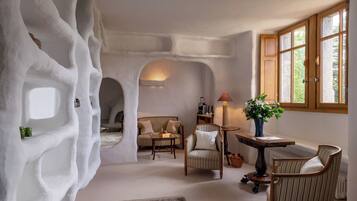 Una habitación con un diseño de pared blanco y único, similar a una cueva, un rincón de descanso acogedor con sofá y sillones, una mesita con una lámpara, y una ventana con vistas a zonas verdes.