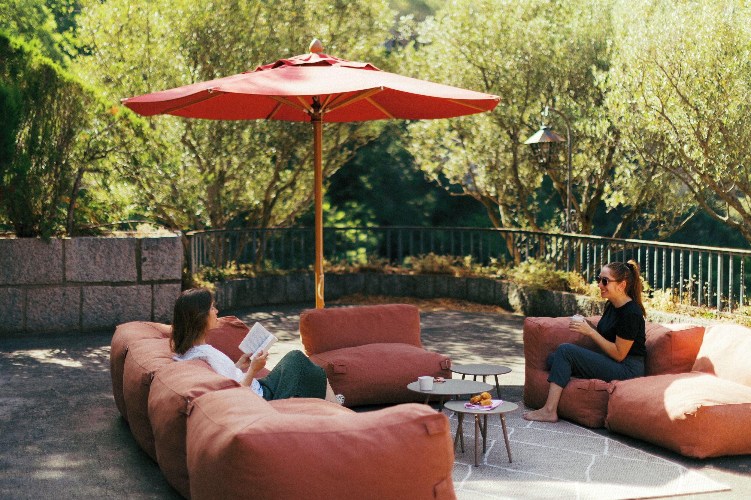 An outdoor seating area with cushioned sofas, a red umbrella, and a small table with a cup and pastries.